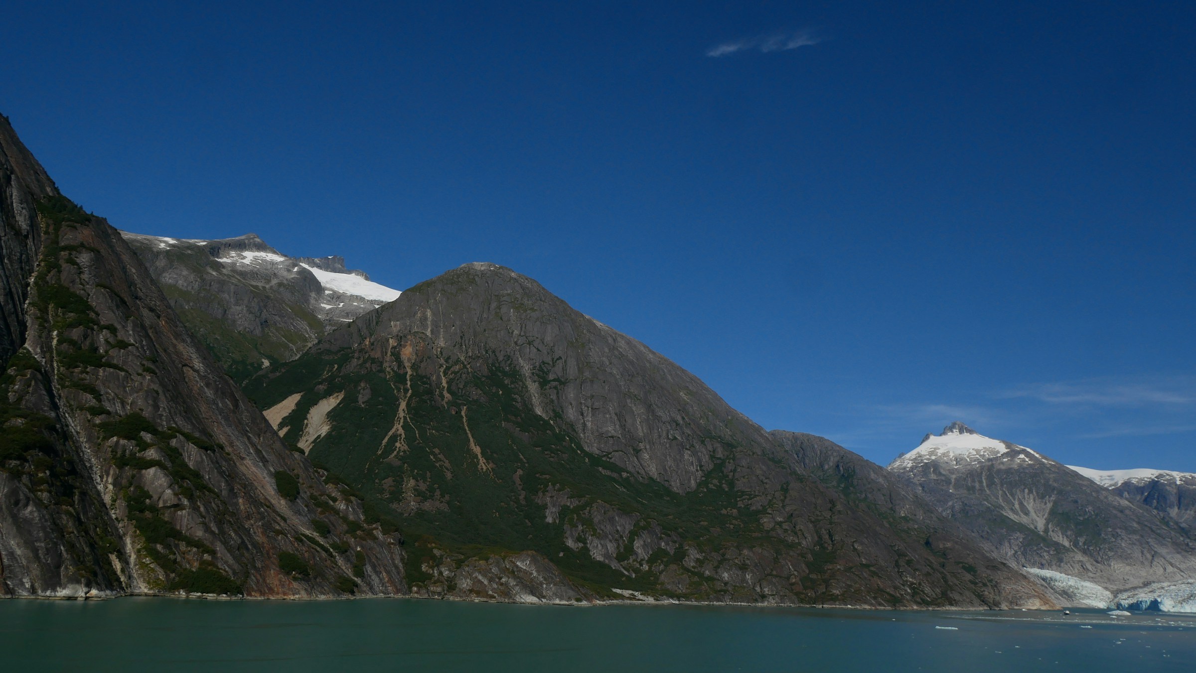 Steep mountains with snow above the calm green water of an Alaskan fjord
